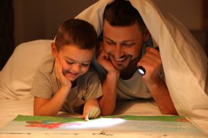 Father and son with flashlight reading book under blanket at home